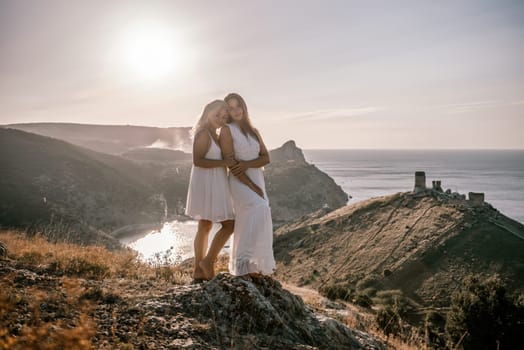 Two young girls are standing on a hillside, one of them wearing a white dress. The sun is shining brightly, creating a warm and inviting atmosphere. The girls seem to be enjoying their time together