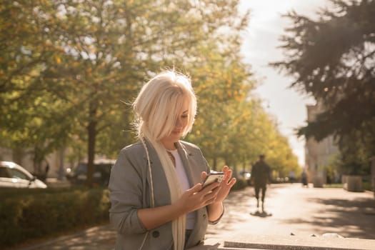 A blonde woman is looking at her cell phone while walking down a sidewalk. The scene is sunny and peaceful, with trees in the background