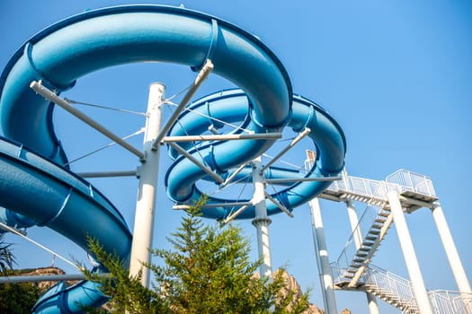A blue waterslide twists and turns against a clear blue sky. The slide is made of metal and has a white frame. A small green tree is visible in the lower left corner.