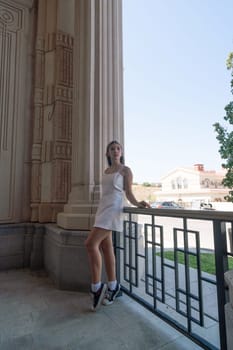 A young woman in a white dress leans on a railing outside a building with columns. She looks towards the right side of the frame.