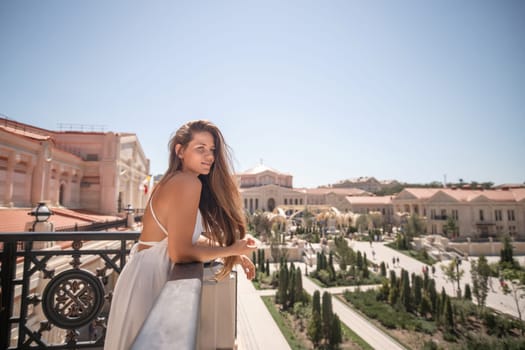 A woman in a white dress stands on a balcony overlooking a sprawling city with gardens and buildings. She looks over her shoulder, gazing out at the view.