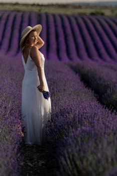 A woman in a white dress stands in a field of purple flowers. She is holding a bouquet of flowers in her hand