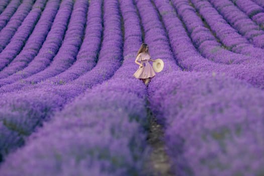 A woman is walking through a field of purple flowers. She is wearing a pink dress and a straw hat. The field is full of lavender flowers, creating a serene and peaceful atmosphere