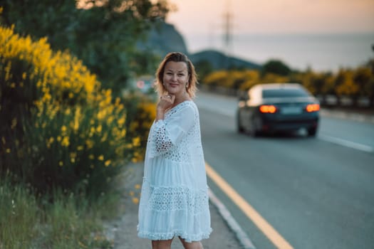A woman in a white dress stands on the side of a road. The road is empty and there is a car in the background. The woman is smiling and looking at the camera