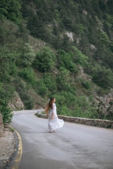 A woman in a white dress is walking down a road. The road is surrounded by trees and the woman is looking ahead