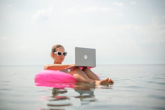 A woman is sitting on a pink inflatable raft in the ocean, using a laptop. Concept of relaxation and leisure, as the woman is enjoying her time in the water while working on her laptop