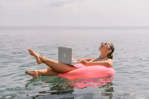 A woman is floating on a pink inflatable raft in the ocean while holding a laptop. Concept of relaxation and leisure, as the woman is enjoying her time in the water while working on her laptop