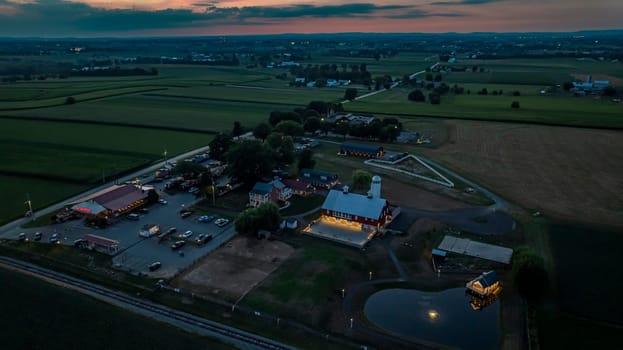Ronks, Pennsylvania, USA, July 19 2024 - Tranquil Farm Scene At Dusk Featuring A Large Red Barn With A White Silo, An Illuminated Stone Courtyard, A Nearby Brick House, Expansive Green Fields, And A Reflective Pond, All Under A Softly Lit Evening Sky.