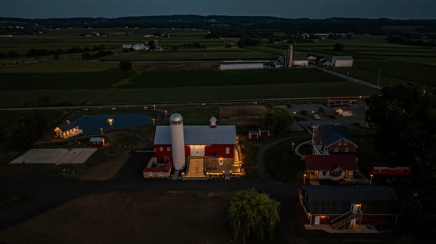 Ronks, Pennsylvania, USA, July 19 2024 - Tranquil Farm Scene At Dusk Featuring A Large Red Barn With A White Silo, An Illuminated Stone Courtyard, A Nearby Brick House, A Reflective Pond, And Expansive Green Fields Under A Softly Lit Evening Sky, Creating A Serene And Picturesque Setting.