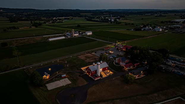 Ronks, Pennsylvania, USA, July 19 2024 - Tranquil Farm Scene At Dusk Featuring A Large Red Barn With A White Silo, An Illuminated Stone Courtyard, A Nearby Brick House, Expansive Green Fields, A Reflective Pond, And A Scenic View Of The Surrounding Countryside Under A Softly Lit Evening Sky.