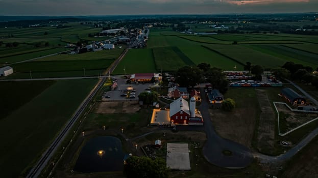Ronks, Pennsylvania, USA, July 19 2024 - Tranquil Farm Scene At Dusk Featuring A Large Red Barn With A White Silo, An Illuminated Stone Courtyard, A Nearby Brick House, Expansive Green Fields, A Reflective Pond, And A Scenic View Of The Surrounding Countryside Under A Softly Lit Evening Sky.