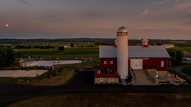 Ronks, Pennsylvania, USA, July 19 2024 - Peaceful Farm Scene At Dusk Featuring A Large Red Barn With A Prominent White Silo, A Stone Pathway, And Surrounding Buildings, All Set Against A Backdrop Of Rolling Green Fields And A Softly Lit Sky.