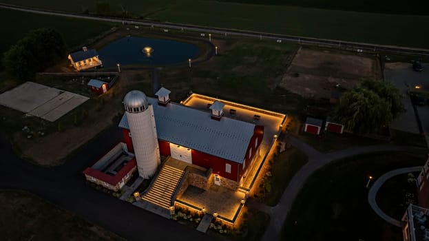 Ronks, Pennsylvania, USA, July 19 2024 - Tranquil Farm Scene At Dusk Featuring A Large Red Barn With A White Silo, An Illuminated Stone Courtyard, A Nearby Cozy Stone House Reflecting In A Pond, Expansive Green Fields, And A Serene Evening Sky
