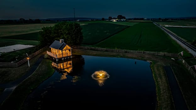 Ronks, Pennsylvania, USA, July 19 2024 - Tranquil Farm Scene At Dusk Featuring A Cozy Stone House Illuminated By Soft Lighting, Reflecting In A Peaceful Pond With A Fountain, Surrounded By Expansive Green Fields