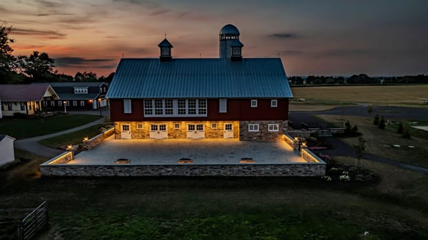 Ronks, Pennsylvania, USA, July 19 2024 - Peaceful Farm Scene At Dusk Featuring A Large Red Barn With A White Silo, An Illuminated Stone Courtyard, Adjacent Buildings, And Expansive Green Fields Under A Beautifully Lit Sunset Sky.