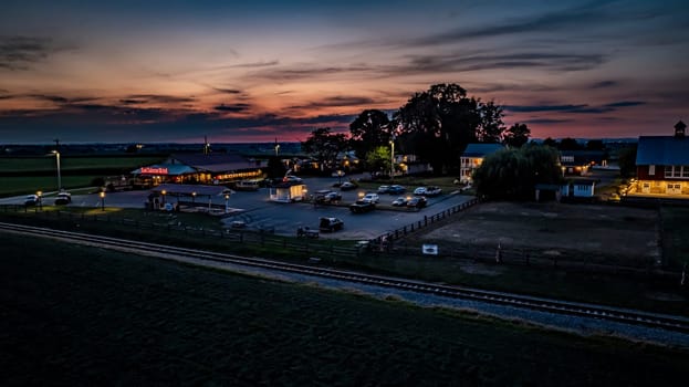 Ronks, Pennsylvania, USA, July 19 2024 - Tranquil Evening Scene Featuring A Farm With A Red Barn, Cozy Buildings, A Parking Lot With Cars, Illuminated By Soft Lighting, A Nearby Railroad Track, And A Vibrant Sunset Sky Creating A Serene Atmosphere.