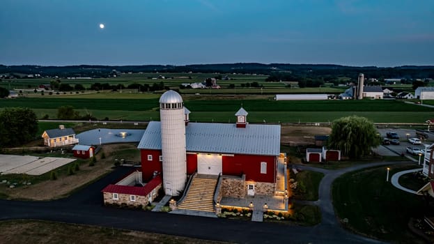 Ronks, Pennsylvania, USA, July 19 2024 - Serene Farm Scene Dusk Featuring Large Red Barn With Silo, Stone Courtyard Illuminated By Soft Lighting, Charming Stone House Reflecting In Pond, Expansive Green Fields, Rising Full Moon.
