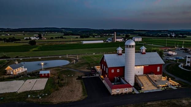 Ronks, Pennsylvania, USA, July 19 2024 - Tranquil Farm Scene At Dusk Featuring A Large Red Barn With A White Silo, An Illuminated Stone Courtyard, Cozy Stone House By Reflective Pond, Expansive Green Fields,