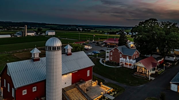 Ronks, Pennsylvania, USA, July 19 2024 - Tranquil Farm Scene At Dusk Featuring A Large Red Barn With A White Silo, An Illuminated Stone Courtyard, A Cozy Stone House By A Reflective Pond, And Expansive Green Fields,