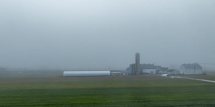 A thick fog envelops a rural setting, revealing silhouettes of farm buildings, a silo, and open fields at dawn.