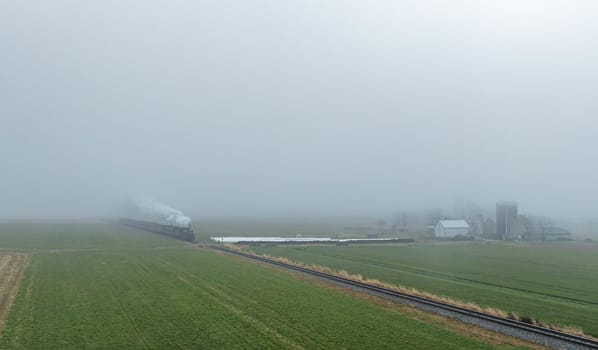 A steam train makes its way through a foggy landscape, surrounded by green fields and distant farm buildings.