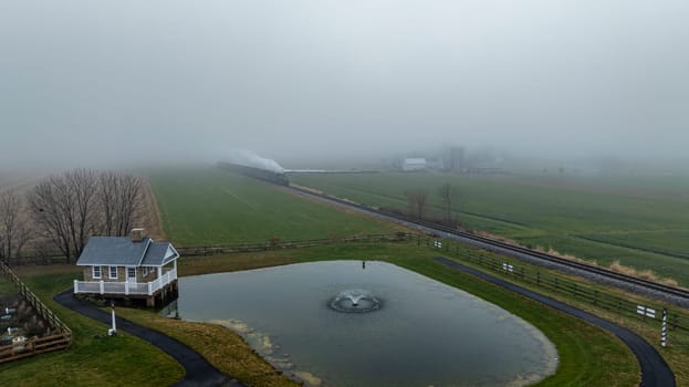 A train travels through a foggy countryside, passing a little house and a pond with a fountain, surrounded by fields.