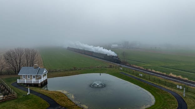 A steam train navigates through fog-covered fields beside a quaint house and a pond, creating a serene atmosphere.