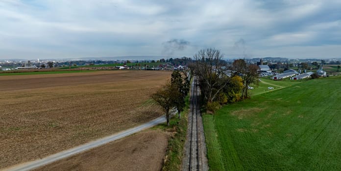 A vast expanse of farmland stretches alongside railway tracks, leading toward a town beneath a cloudy sky.