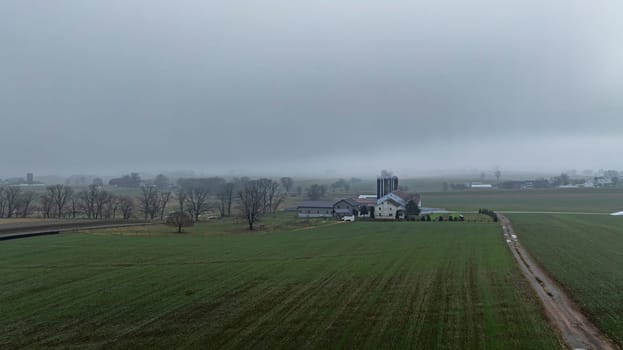 A rural farmstead is enveloped in fog, surrounded by expansive green fields and distant structures on a calm morning.