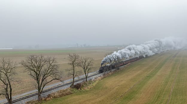 A steam train puffs clouds of smoke while traveling alongside fog-covered fields under an overcast sky.