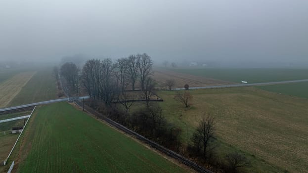 A misty morning covers a rural area, highlighting scattered trees and expansive fields, with a road partially visible.