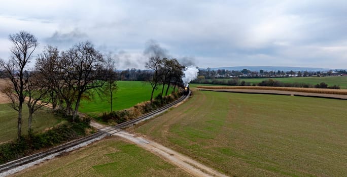 A steam train chugs along winding tracks, billowing smoke, surrounded by green fields and distant hills.