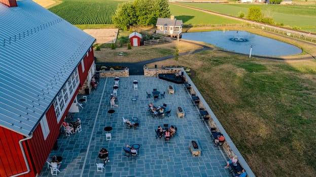 Ronks, Pennsylvania, USA, July 11 2024 - Outdoor Patio With Stone Pavers And Seating Area, Overlooking A Small Pond And Scenic Farmland, With People Relaxing And Enjoying The View Under A Clear Blue Sky.