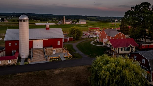 Ronks, Pennsylvania, USA, July 19 2024 - Peaceful Farm Scene At Dusk Featuring A Large Red Barn With A Prominent White Silo, A Stone Pathway, And Surrounding Buildings, All Set Against A Backdrop Of Rolling Green Fields And A Softly Lit Sky.