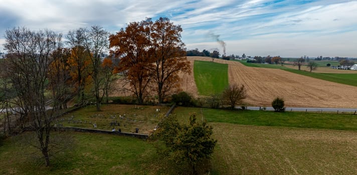 A scenic view of autumn-hued trees near harvested fields, and an old Cemetery, with smoke billowing in the distance under a cloudy sky.