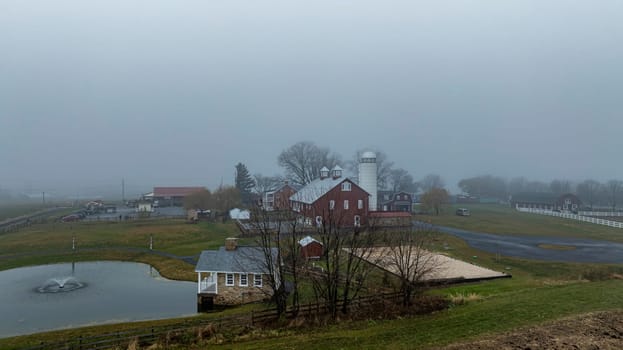 A red barn and small house sit by a pond, surrounded by grass, under a thick layer of fog on an overcast day.
