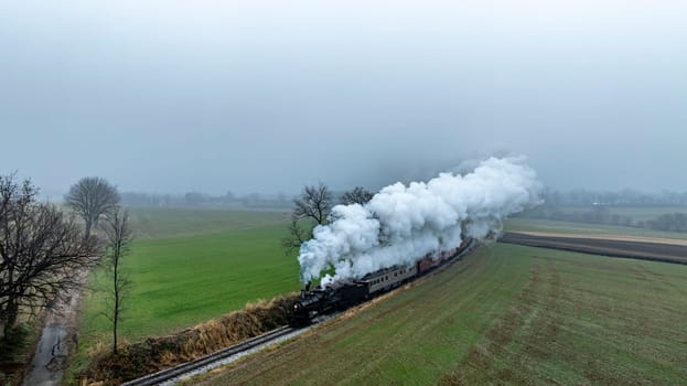 A steam train chugs through green fields, releasing clouds of smoke into the misty air as it makes its way along the tracks.