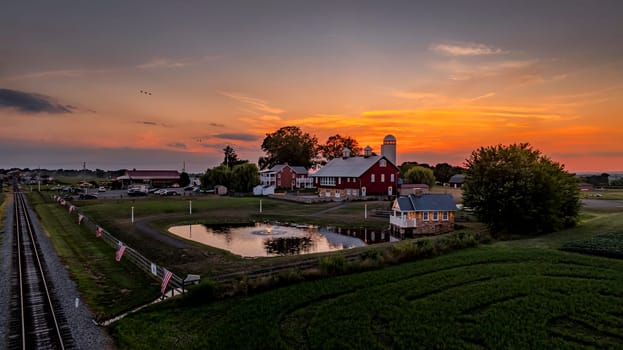 Ronks, Pennsylvania, USA, July 19 2024 - Tranquil Farm Scene At Sunset Features Red Barn, Silos, Pond, Surrounding Green Fields, With Train Tracks In The Foreground Dramatic, Colorful Sky Illuminating The Horizon.