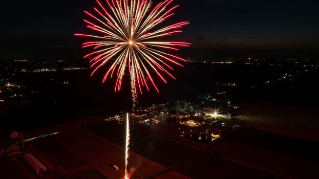 Bright Red Fireworks Explode In The Night Sky, Illuminating A Rural Fairground Below With Bustling Activity, Surrounded By Expansive Dark Fields And Distant City Lights On The Horizon.