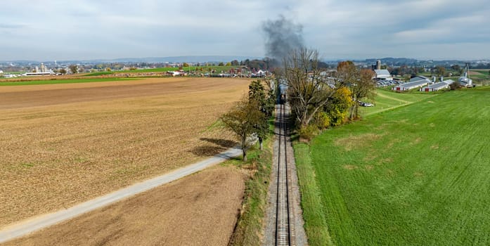 A train moves along tracks surrounded by fields and trees, with a rural town visible in the background under gray skies.