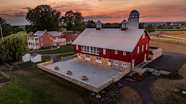 Ronks, Pennsylvania, USA, July 19 2024 - Tranquil Farm Scene At Sunset Features A Large Red Barn With A Stone Foundation, A Spacious Stone Courtyard, A Nearby Brick House, And A Colorful Sky, Creating A Peaceful And Picturesque Setting.