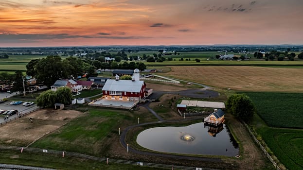 Ronks, Pennsylvania, USA, July 19 2024 - Scenic Farm Landscape At Dusk Showcasing A Large Red Barn With A White Silo, A Stone Courtyard, A Charming Stone House By A Pond, And Expansive Green Fields Stretching Into The Distance