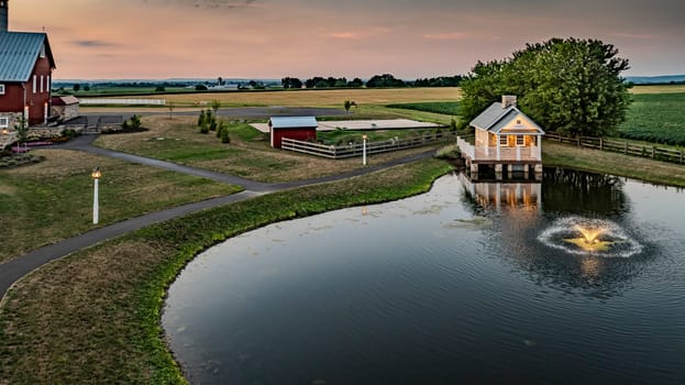 Ronks, Pennsylvania, USA, July 19 2024 - Charming Farm Scene At Dusk Featuring A Small Stone House With A White Porch Reflecting In A Pond, Surrounded By Green Fields, Fenced Pastures, And A Softly Lit Evening Sky, Creating A Tranquil Setting.