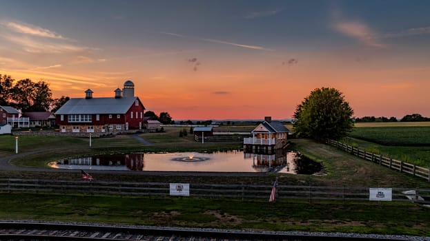 Ronks, Pennsylvania, USA, July 19 2024 - Red Barn With Silver Roof And Cupolas At Sunset, Reflected In A Pond With A Fountain, Surrounded By Open Fields, Fencing, Trees, And A Small House Under A Vibrant Sky.