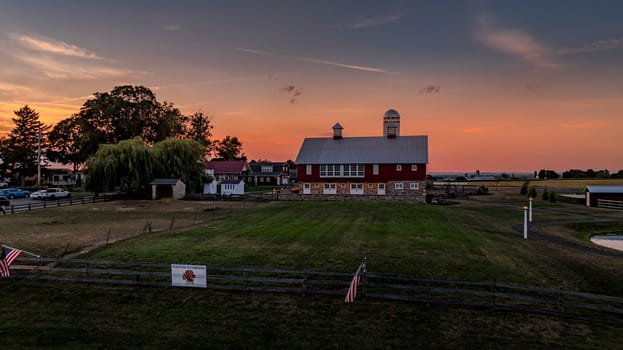 Ronks, Pennsylvania, USA, July 19 2024 - Large Red Barn With Silver Roof And Twin Cupolas At Sunset, Surrounded By Open Fields, A Weeping Willow, Fencing, And Other Farm Structures Under A Pink And Orange Sky.