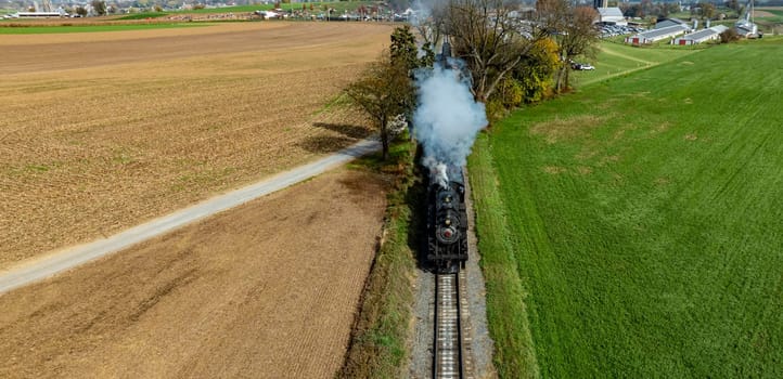 A vintage steam train chugs along grassy fields and farmland, surrounded by vibrant autumn colors.