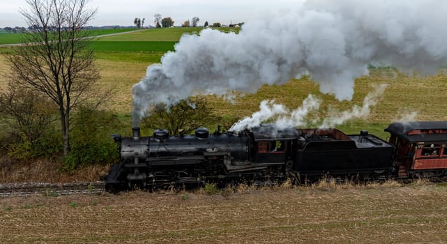 A vintage steam locomotive travels on tracks through a field, releasing thick clouds of smoke into the overcast sky.