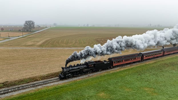 A steam locomotive chugs along railway tracks, releasing clouds of smoke as it passes through expansive farmland under a grey sky.