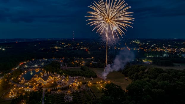 White Fireworks Burst In The Night Sky Over A Brightly Lit Fairground With Illuminated Buildings, Surrounded By Lush Greenery And A Distant Town Glowing With City Lights In The Horizon.