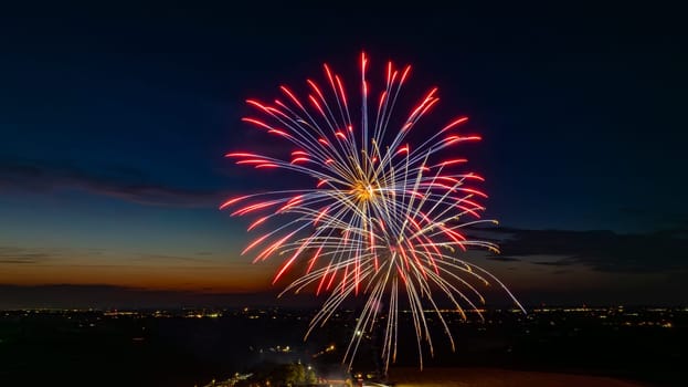 Red And Blue Fireworks Exploding In The Night Sky, Forming Brilliant Bursts Over The Distant City Lights And A Gathered Crowd, With The Twilight Horizon Enhancing The Festive Atmosphere.